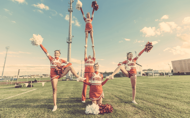 Cheerleaders perform stunts and tumbling on a vibrant, crowded gym floor. A pyramid of athletes, with arms outstretched, supports a flyer mid-air